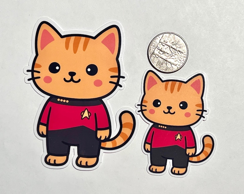 May include: Two orange tabby cat stickers, one large and one small, wearing red and black uniforms with a gold insignia. A US quarter is included for size comparison. The cats have large black eyes and pink cheeks.