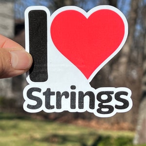 May include: A white sticker with a black 'I' and a red heart, and the word 'Strings' in black below.
