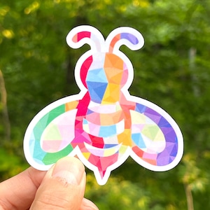 May include: A colorful geometric bee sticker with a white outline. The bee is made up of various shades of pink, red, orange, yellow, green, blue, and purple.