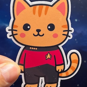 May include: A sticker featuring a cartoon cat in a Starfleet uniform. The cat has orange fur with darker stripes, pink ears and cheeks, and black eyes. The uniform is red and black with a gold insignia.