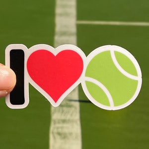 May include: A sticker with the words "I ❤️ Tennis". The "I" is black, the heart is red, and the tennis ball is green with white details. The background is a green tennis court.