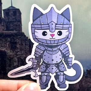 May include: A sticker featuring a cartoon cat in medieval armor, holding a sword. The cat has a white face, pink nose, and black eyes. The armor is a shade of gray, and the background shows a castle.