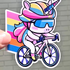 May include: A colorful unicorn wearing a helmet and sunglasses rides a pink bicycle with white wheels. The unicorn has a rainbow mane and tail.