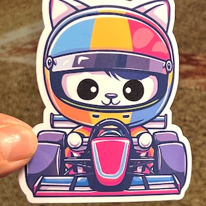 May include: A cartoon sticker of a cat wearing a colorful racing helmet and driving a race car. The cat has big eyes and a cute expression. The car is purple and pink with a red front.