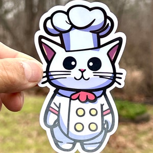 A white cat wearing a chef's hat and uniform with a red bow tie and four yellow buttons. The cat is smiling and has big black eyes. he waterproof vinyl decal features a clean white border for decorating water bottles, car windows, and laptops.