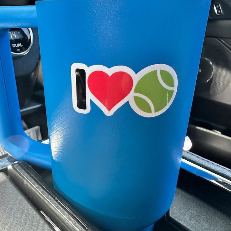 May include: A blue travel mug with a handle, featuring a sticker that says "I ❤️ Tennis." The sticker has a red heart and a green tennis ball. The mug is in a car cup holder.