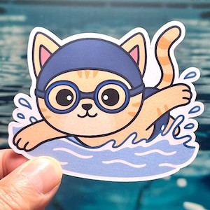 May include: A sticker featuring a cartoon cat swimming in a pool. The cat is orange and white, wearing a blue swim cap and goggles. Water splashes surround the cat, creating a playful and dynamic image. The sticker is held by a person.