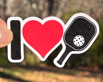Pode incluir: Um autocolante com as palavras "I ❤️" seguidas de um coração vermelho e uma raquete de pickleball preta com uma bola branca. O autocolante tem uma borda branca e é segurado contra um fundo desfocado ao ar livre.