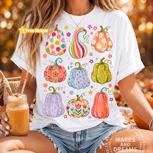 May include: A white t-shirt featuring a colorful graphic print of nine stylized pumpkins in various patterns, including polka dots, floral motifs, and stripes, interspersed with small flowers and stars. The design is vibrant and multi-colored.