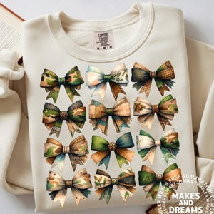 May include: Cream-colored sweatshirt featuring a grid of twelve watercolor bows in various patterns and colors, including camouflage, floral, and plaid. The bows have a rustic, vintage aesthetic. The text "MAKES AND DREAMS" is at the bottom.