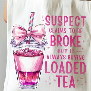 May include: A white tote bag with a pink and white illustration of a cup of iced tea with a pink bow and straw. The text "SUSPECT CLAIMS TO BE BROKE BUT IS ALWAYS BUYING LOADED TEA" is written in pink letters.