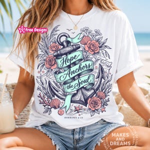May include: A white short-sleeve t-shirt featuring a graphic illustration of a large anchor wrapped in a light blue ribbon with the text "Hope Anchors the Soul" and "HEBREWS 6:19". The design includes pink roses, seashells, and coral.