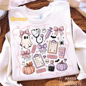 May include: A cream-colored crewneck sweatshirt featuring a graphic print of medical and Halloween-themed icons in pastel pink and purple. The design includes ghosts, a stethoscope, a syringe, clipboards, bows, pumpkins, and bandages.
