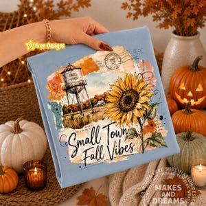 May include: A light blue sweatshirt featuring a graphic print of a water tower, a large yellow sunflower, and the text Small Town Fall Vibes. The design includes watercolor-style patches in orange and brown, along with a vintage postmark stamp.