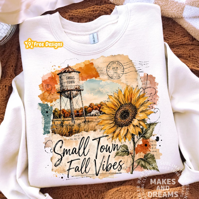 May include: A cream-colored crewneck sweatshirt featuring a graphic print of a water tower labeled Small Town, a large yellow sunflower, and the text Small Town Fall Vibes. The design includes vintage-style postal stamps and watercolor textures.