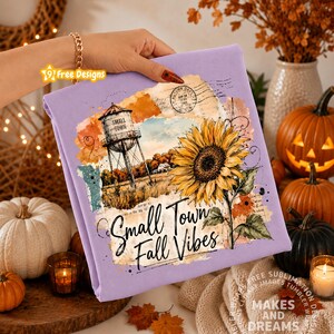 May include: A light purple sweatshirt featuring a graphic print of a water tower, a large yellow sunflower, and the text "Small Town Fall Vibes" in a black script font, layered over a vintage-style collage of paper textures and postmarks.