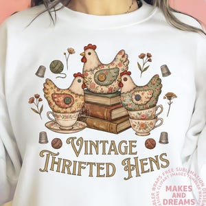 May include: White sweatshirt featuring a vintage-style graphic. The design includes three decorative hens atop books and teacups, with floral and button details. The text "VINTAGE THRIFTED HENS" is displayed in a decorative font.