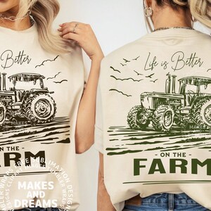 May include: Cream-colored t-shirt featuring a detailed black and white tractor graphic. The text "Life is Better" is above the tractor, with "- ON THE - FARM" below. The shirt design includes birds and is from "MAKES AND DREAMS".
