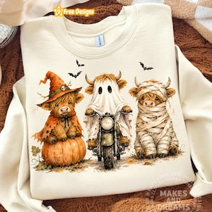Op de afbeelding: Een crèmekleurig sweatshirt met ronde hals en een afbeelding van drie Highland-koeien verkleed voor Halloween. Eén koe draagt een heksenhoed op een pompoen, één rijdt op een motor als spook en één is ingewikkeld als een mummie.