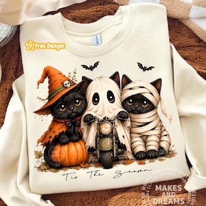 May include: A cream-colored sweatshirt featuring a graphic of three black kittens dressed for Halloween. One kitten wears an orange witch hat on a pumpkin, one is a ghost riding a motorcycle, and one is a mummy. The text reads 'Tis The Season'.