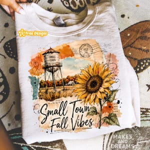 May include: A cream-colored crewneck sweatshirt featuring a graphic print of a water tower, a large sunflower, and the text "Small Town Fall Vibes" with a vintage postmark stamp. The design includes watercolor-style orange and brown accents.