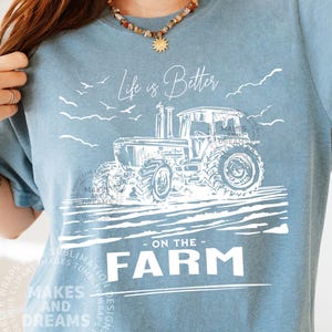 May include: A light blue t-shirt with a white graphic of a tractor and the text "Life is Better on the Farm." The design includes birds and clouds. The shirt is from the "Makes and Dreams" collection.