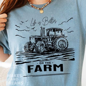 May include: A light blue t-shirt featuring a black illustration of a tractor on a farm. The text "Life is Better - ON THE - FARM" is printed below the tractor. The shirt has a crew neck and short sleeves.