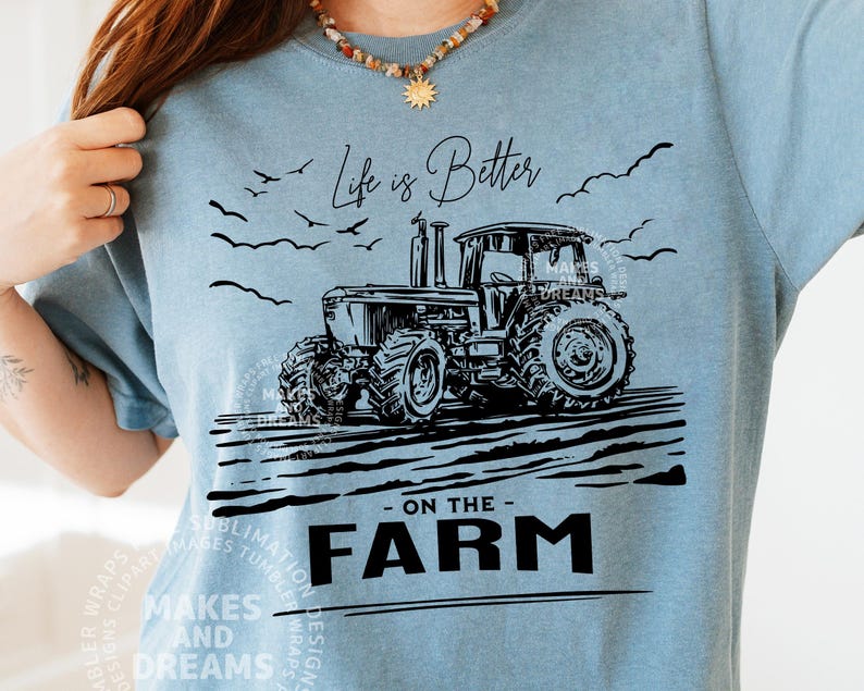 May include: A light blue t-shirt featuring a black illustration of a tractor on a farm. The text "Life is Better - ON THE - FARM" is printed below the tractor. The shirt has a crew neck and short sleeves.