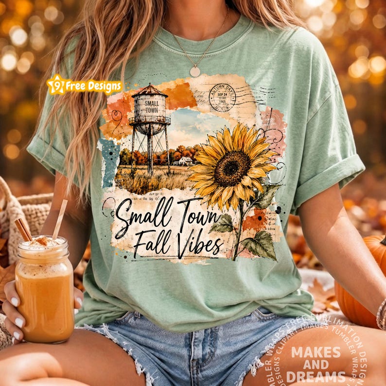 May include: A light green t-shirt featuring a graphic print of a water tower, a large yellow sunflower, and the text "Small Town Fall Vibes" in a script font, layered over a background of vintage paper textures and postal stamps.