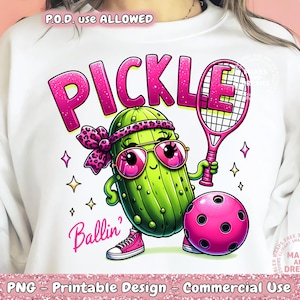 Pode incluir: Uma camiseta branca com um picles de desenho animado rosa e verde segurando uma raquete de tênis rosa e uma bola rosa e preta. O texto "PICKLE Ballin'" está impresso na camiseta.