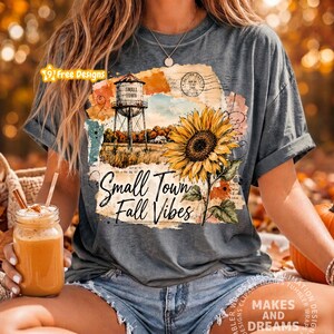 May include: A charcoal gray t-shirt featuring a graphic print of a sunflower, a water tower, and the text Small Town Fall Vibes. The design includes vintage-style postal stamps and script elements layered over orange and cream watercolor textures.