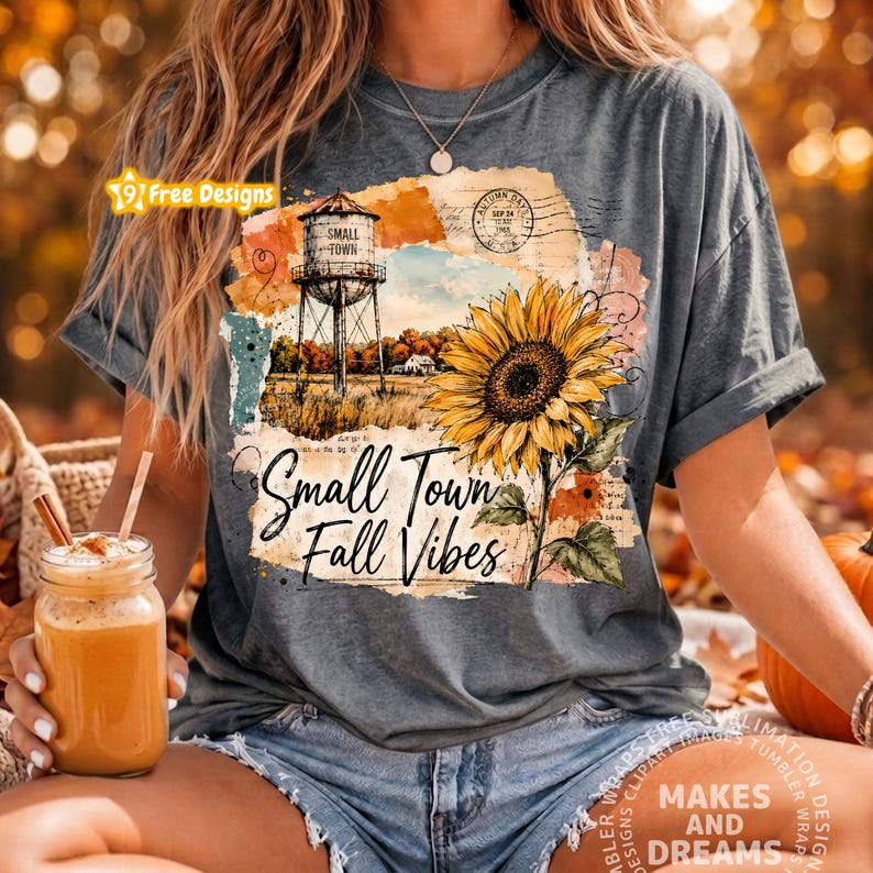 May include: A charcoal gray t-shirt featuring a graphic print of a sunflower, a water tower, and the text Small Town Fall Vibes. The design includes vintage-style postal stamps and script elements layered over orange and cream watercolor textures.