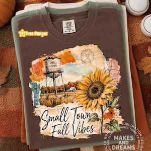 May include: A brown short-sleeve t-shirt featuring a graphic print of a water tower, a large yellow sunflower, and a postmark stamp. The text reads Small Town and Small Town Fall Vibes. The shirt has a Comfort Colors brand label at the neck.