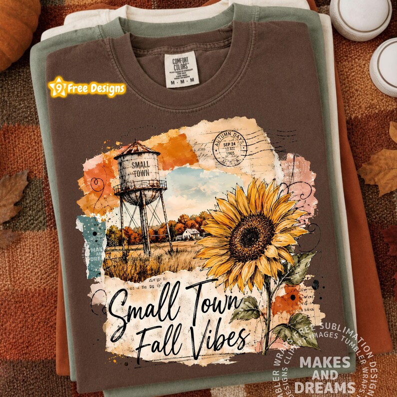 May include: A brown short-sleeve t-shirt featuring a graphic print of a water tower, a large yellow sunflower, and a postmark stamp. The text reads Small Town and Small Town Fall Vibes. The shirt has a Comfort Colors brand label at the neck.