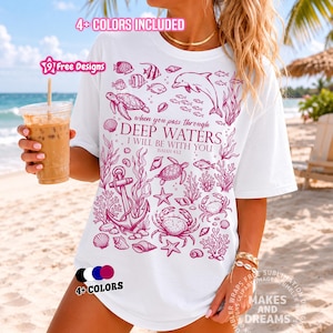 May include: A white t-shirt featuring a magenta graphic print of marine life including a dolphin, sea turtles, crabs, shells, and an anchor. The text reads: when you pass through DEEP WATERS I WILL BE WITH YOU ISAIAH 43:2. The design is centered.