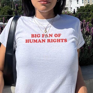 May include: White t-shirt with the red text "BIG FAN OF HUMAN RIGHTS". The short-sleeved shirt is likely cotton. The person is wearing sunglasses and a necklace. The background includes greenery and buildings.