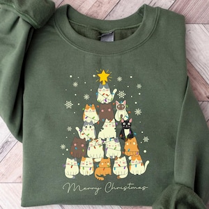 May include: A green sweatshirt featuring a Christmas tree design made of cats. The cats are various colors and are decorated with lights and a star on top. The words "Merry Christmas" are written in a script font below the tree.