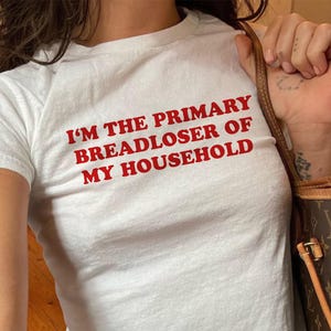 May include: A white short-sleeved t-shirt featuring the red text I'M THE PRIMARY BREADLOSER OF MY HOUSEHOLD printed on the chest. The shirt is paired with blue denim jeans and a brown patterned shoulder bag with gold-toned hardware accents.