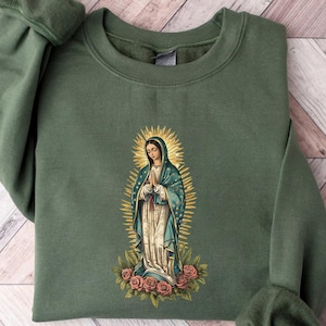 May include: An olive green sweatshirt featuring an embroidered image of the Virgin Mary with a golden halo, blue robe, and roses at the bottom. The sweatshirt has long sleeves with ribbed cuffs and a crew neck.