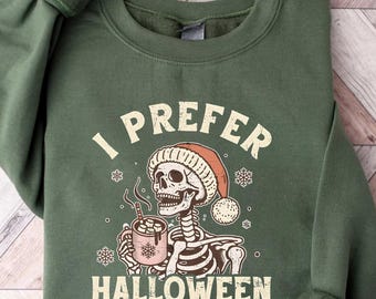 May include: A dark green sweatshirt with the text "I PREFER HALLOWEEN" in a vintage style. The graphic features a skeleton wearing a Santa hat and holding a mug. The sweatshirt is folded on a wooden surface.