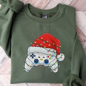 Puede incluir: Una sudadera verde oliva con un diseño festivo. El gráfico representa un mando de videojuegos con un gorro de Papá Noel adornado con luces navideñas. La sudadera está colocada sobre una superficie clara con vetas de madera.
