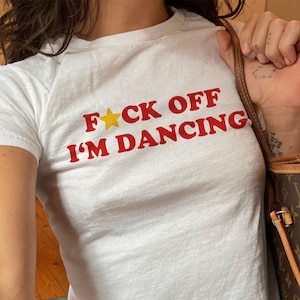 Puede incluir: Una camiseta blanca de manga corta con el texto rojo F*CK OFF I'M DANCING, donde una estrella amarilla sustituye a la letra U. La prenda la lleva un modelo de 54 kg en talla XS, combinada con vaqueros azules y un bolso marrón estampado.