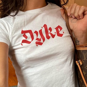 Peut inclure: T-shirt blanc avec le mot "Dyke" en lettres gothiques rouges. Le t-shirt est à manches courtes et porté avec un jean bleu. Un sac à main marron avec un motif monogramme est visible.