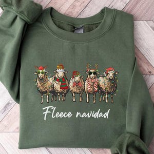 May include: An olive green sweatshirt featuring a graphic of five sheep wearing festive holiday accessories, including hats and scarves. The text "Fleece navidad" is printed below the sheep. The sweatshirt is made of a soft material.