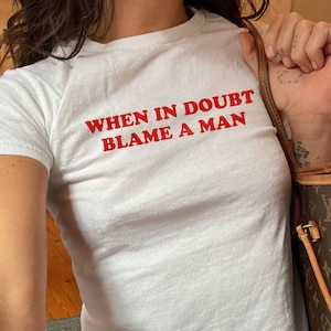 May include: White t-shirt with the red text "WHEN IN DOUBT BLAME A MAN." The shirt is short-sleeved and appears to be made of cotton. The model is wearing blue jeans and carrying a brown handbag.