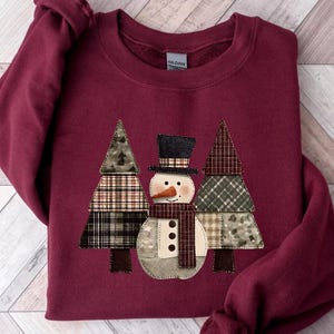 May include: Burgundy crew neck sweatshirt with a snowman and two Christmas trees appliquéd on the front. The snowman has a black top hat, carrot nose, and scarf. The trees and snowman are made of patchwork fabric.