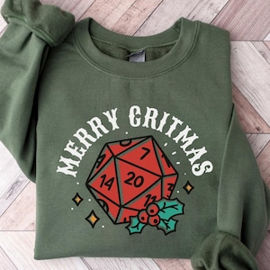 Puede incluir: Sudadera verde con el texto "MERRY GRITMAS" sobre un dado D20 rojo con números blancos y acebo. La sudadera está doblada sobre una superficie de vetas de madera clara.