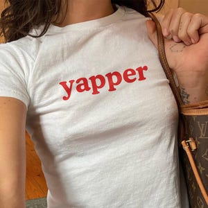 May include: White crew neck t-shirt with the word "yapper" printed in red. The t-shirt is worn with blue denim jeans. A brown handbag with a monogram pattern is visible.