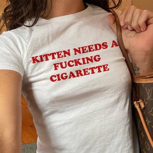 Op de afbeelding: Een wit T-shirt met korte mouwen en de rode tekst KITTEN NEEDS A FUCKING CIGARETTE op de borst. Het shirt heeft een ronde hals en een aansluitende pasvorm, gecombineerd met een bruine tas met monogram en lederen schouderbanden.