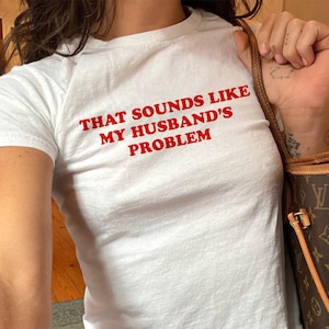 May include: White t-shirt with the red text "THAT SOUNDS LIKE MY HUSBAND'S PROBLEM." The shirt is paired with blue jeans. A brown and tan handbag is visible in the image.